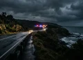 A dark, moody Australian highway at night with emergency vehicle lights blurred in the distance.