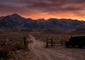 A silhouetted vehicle near a ranch gate at sunset in a mountainous landscape.