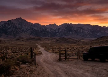 A silhouetted vehicle near a ranch gate at sunset in a mountainous landscape.