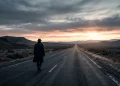 A solitary muscular figure walks down a long highway at dawn.