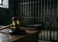 A gavel on a wooden desk next to out-of-focus jail bars in a dark room.