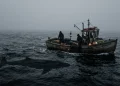 A small fishing boat on dark, foggy waters with a mysterious shape lurking beneath the surface.