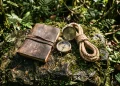 A conceptual arrangement of treasure hunting equipment including a leather journal and a brass compass on a jungle rock.